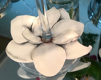 May include: A clear glass wine glass with a white magnolia flower design. The flower is printed on a paper-like material and sits atop the glass. The stem of the glass is visible, and the background is a reflective surface.