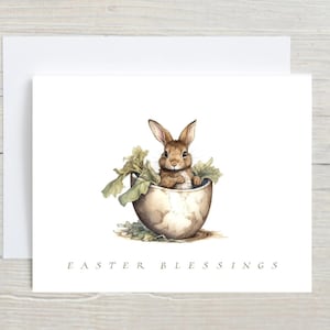 May include: Watercolor Easter card featuring a brown bunny emerging from a cracked eggshell, surrounded by green leaves. The card has the text "EASTER BLESSINGS" at the bottom, and is accompanied by a white envelope.