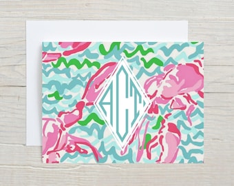 May include: A stationery set featuring a patterned design with pink floral elements, teal and green wavy lines, and a white diamond-shaped monogram. The set includes a card and a white envelope.