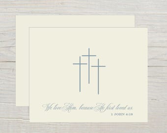 May include: Off-white stationery set featuring three light blue crosses. The phrase "We love Him, because He first loved us. 1 John 4:19" is printed in a script font below the crosses. Includes an envelope.