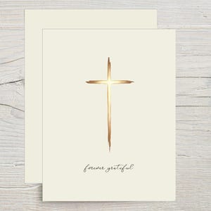 May include: A cream-colored greeting card featuring a centered, vertical cross rendered in a metallic copper brushstroke style. Below the cross, the text "forever grateful" is printed in a delicate, black, cursive script font.