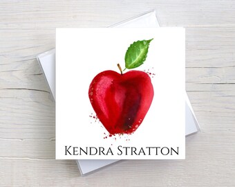 May include: A square white card featuring a watercolor illustration of a red apple with a green leaf. The apple has a heart-like shape with red paint splatters. The name "Kendra Stratton" is printed in black below the apple.