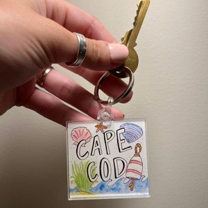 May include: A clear acrylic keychain with a hand-drawn illustration of a seashell, starfish, and a buoy. The text "CAPE COD" is written in black lettering on the keychain.