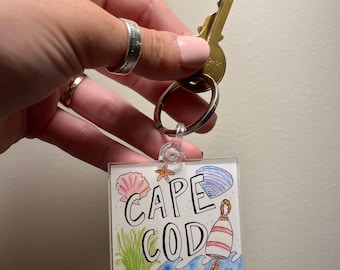 May include: A clear acrylic keychain with a hand-drawn illustration of a seashell, starfish, and a buoy. The text "CAPE COD" is written in black lettering on the keychain.