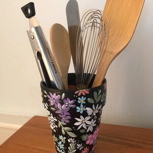 May include: A black flower pot with a floral design in shades of pink, purple, blue, and white. The pot is filled with kitchen utensils, including a wooden spoon, a whisk, a spatula, and tongs.