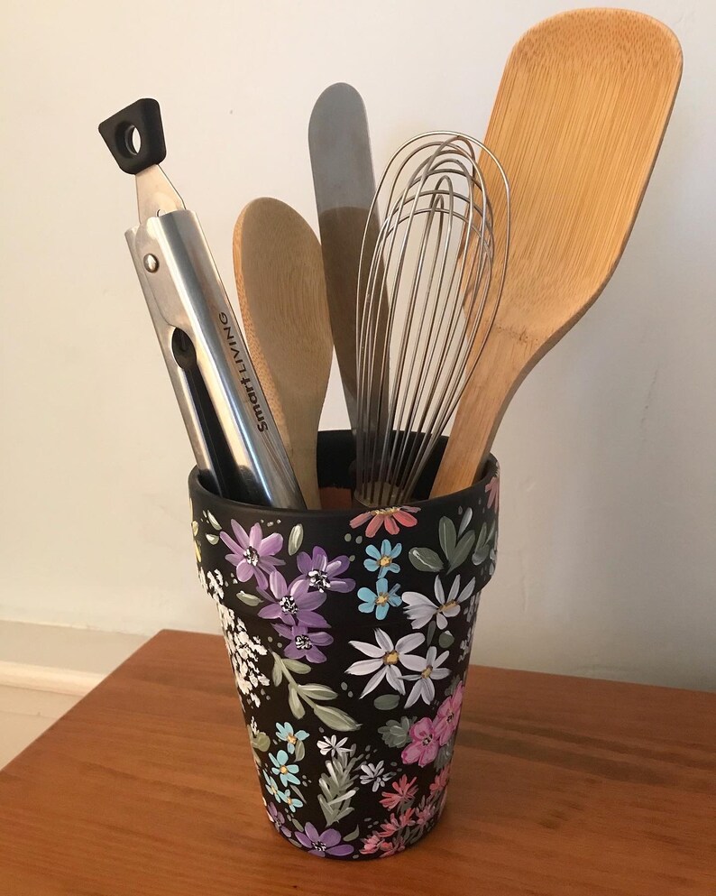 May include: A black flower pot with a floral design in shades of pink, purple, blue, and white. The pot is filled with kitchen utensils, including a wooden spoon, a whisk, a spatula, and tongs.