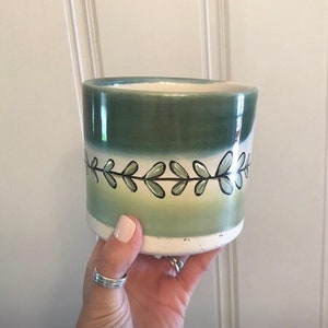 May include: A ceramic planter with a green and white gradient design. The planter features a hand-painted leaf pattern in black and green. The interior is a light blue color. The planter is held by a person.