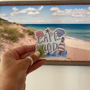 May include: A colorful sticker with the words "CAPE COD" and beach-themed illustrations. The sticker features a seashell, starfish, and a buoy. The background shows a beach scene with blue water and a cloudy sky.