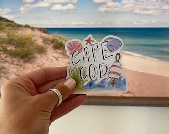 May include: A colorful sticker with the words "CAPE COD" and beach-themed illustrations. The sticker features a seashell, starfish, and a buoy. The background shows a beach scene with blue water and a cloudy sky.