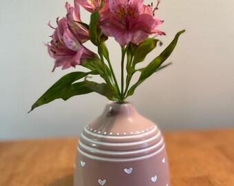 May include: A small, light pink ceramic vase with white painted hearts and dots. The vase is filled with pink alstroemeria flowers.