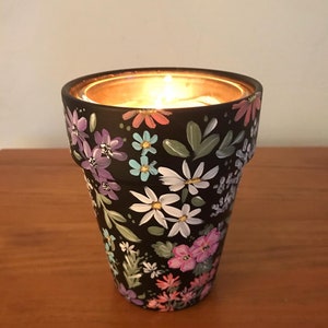 May include: A black ceramic candle holder with a floral design painted in shades of pink, purple, blue, and white. The candle holder has a small opening at the top for a tea light candle.