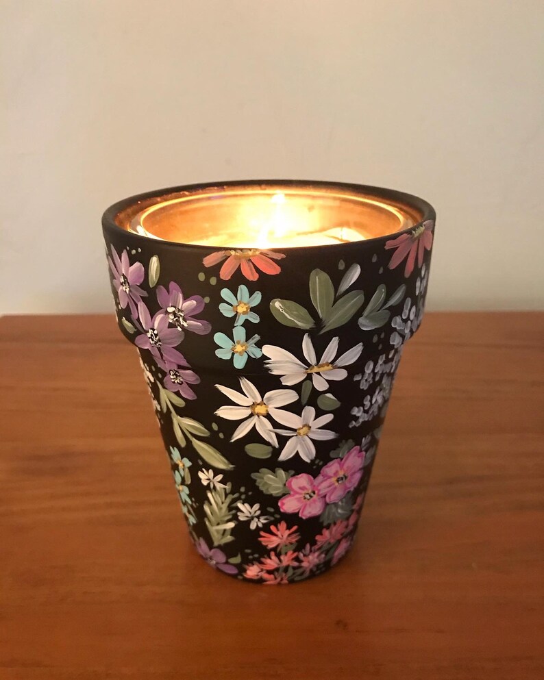 May include: A black ceramic candle holder with a floral design painted in shades of pink, purple, blue, and white. The candle holder has a small opening at the top for a tea light candle.