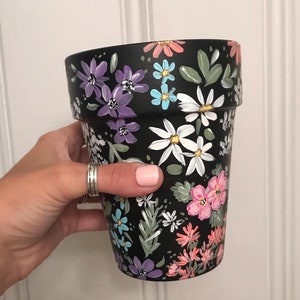 May include: A black terracotta flower pot with a hand-painted floral design. The design features white, pink, purple, blue, and green flowers on a black background.
