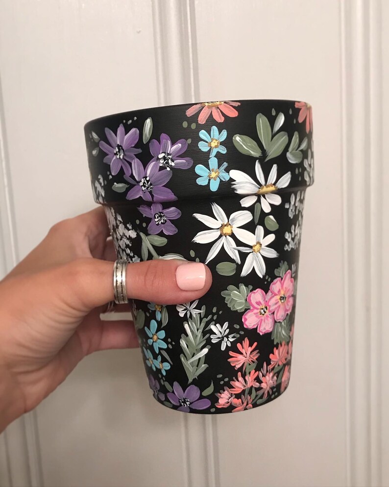 May include: A black terracotta flower pot with a hand-painted floral design. The design features white, pink, purple, blue, and green flowers on a black background.