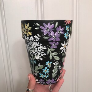 May include: Black terracotta flower pot with a hand-painted floral design. The design features a variety of flowers in shades of purple, blue, yellow, and white.