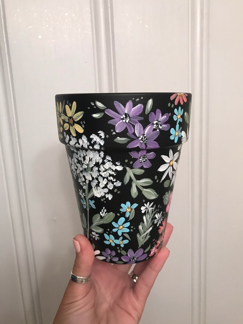 May include: Black terracotta flower pot with a hand-painted floral design. The design features a variety of flowers in shades of purple, blue, yellow, and white.