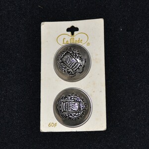 May include: Two silver-toned buttons with a raised design featuring a building and foliage. The buttons are on a white card with the text "La Mode" and "60¢".