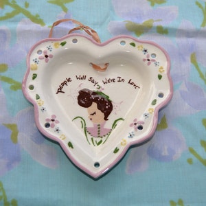 May include: A white heart-shaped ceramic dish with a pink rim. The dish has a painted design of a woman with brown hair and a floral pattern. The text "People will say we're in love" is written on the dish.