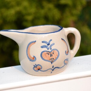 Op de afbeelding: Een kleine witte keramische roomkan met een blauw en oranje bloemenmotief. De roomkan heeft een handvat en een tuit.