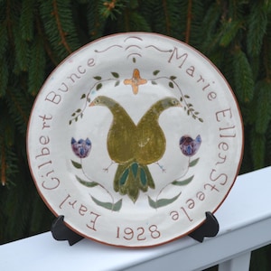 May include: A round ceramic plate with a cream-colored glaze featuring a central illustration of two conjoined olive-green birds. The text "Earl Gilbert Bunce" and "Marie Ellen Steele" is inscribed along the rim with the year "1928" at the bottom.