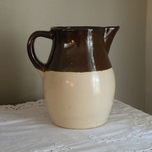 May include: A brown and white ceramic jug with a handle. The jug is glazed in a brown colour on the top half and a white colour on the bottom half.