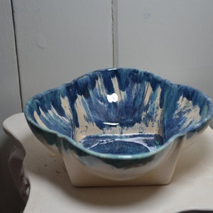 May include: A white ceramic bowl with a blue and green glaze. The bowl has a square shape with rounded edges and a wavy rim.