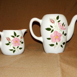 May include: A white ceramic teapot and creamer set with a pink rose floral design. The teapot has a handle and a spout, while the creamer has a handle.