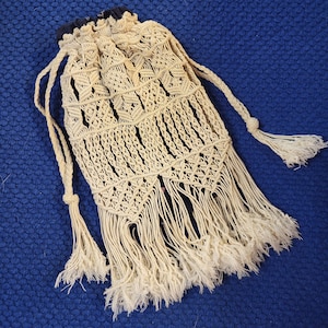 May include: A beige macrame bag with fringe detailing. The bag has a drawstring closure and is made of natural fibers.