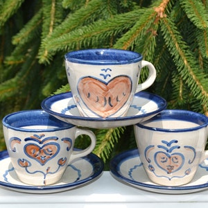 Puede incluir: Tres tazas de té de cerámica y platillos con ribete azul y blanco y un diseño de corazón marrón. Las tazas están apiladas una encima de la otra.