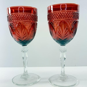 May include: Two matching wine glasses featuring deep red bowls with a textured diamond pattern on the upper section and a leaf-like motif below. Each glass has a clear, faceted stem and a smooth, circular clear glass base for stability.