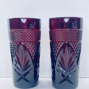 May include: Two tall, cylindrical drinking glasses made of deep red glass. Each glass features a textured upper band with a diamond-cut pattern and a lower section decorated with large, embossed leaf-like motifs and starburst designs.