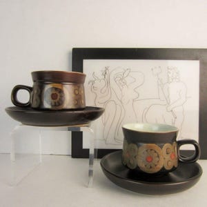 May include: Two brown ceramic teacups and saucers with a floral design. The cups have a dark brown exterior with a lighter brown and red floral pattern. The saucers are dark brown. One cup is on a clear acrylic stand.