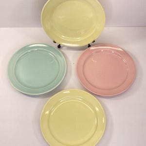 May include: Four pastel-colored ceramic plates. The plates are in shades of yellow, mint green, and pink. The plates are round with a slightly raised rim. One yellow plate is on a black metal stand.