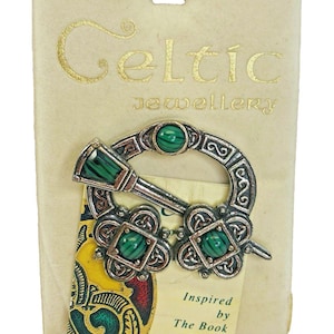 May include: Celtic jewelry brooch with green malachite stones and silver-tone metalwork. The brooch is on a cream-colored card with the words "Celtic Jewellery" in gold lettering. The card also reads "Inspired by The Book of Kells."