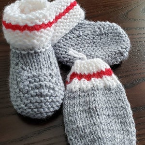 Op de afbeelding: Een paar grijze gebreide babyschoentjes met witte en rode bies. De schoentjes zijn gemaakt met een eenvoudige breisteek en hebben een puntige teen.
