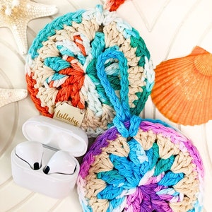 以下が含まれることがあります： 青、ピンク、緑、オレンジの色合いでカラフルな渦巻き模様が施された、丸い形のクロシェ編みエアポッドケースが2つ。それぞれにベージュのループハンドルと「Lullaby」と書かれた木製のタグが付いています。