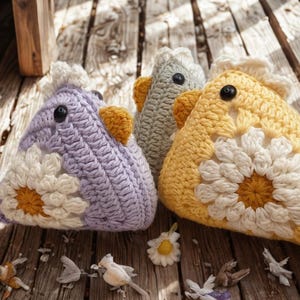 May include: Three crocheted chicken-shaped items in lavender, gray, and yellow, each with a white daisy and a yellow beak. Small decorative birds and flower petals are scattered on the wooden surface.
