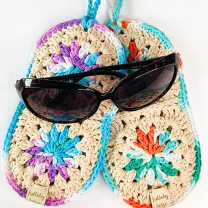 May include: Two crocheted pot holders in a beige, white, pink, blue, and orange color scheme. The pot holders have a circular design with a flower-like pattern in the center. The pot holders are hanging from a loop and have a wooden tag that says "Lullaby Lodge".