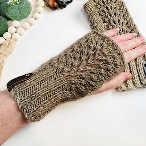 May include: A pair of knitted fingerless gloves in a neutral brown color. The gloves feature a detailed leaf-like pattern and a leather strap with a gold-colored button. The gloves are designed to keep hands warm while leaving fingers free.