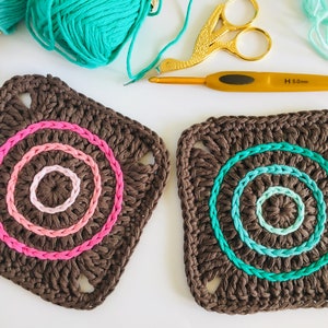 May include: Two brown crochet squares with colorful yarn circles in the center. The square on the left has pink, light pink, and white yarn circles. The square on the right has teal, light teal, and white yarn circles.