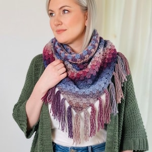 May include: A triangular crochet shawl featuring a gradient of colors including deep purple, magenta, and soft blue. The shawl is constructed with a textured stitch pattern and is finished with long, matching yarn fringe along the bottom edges.