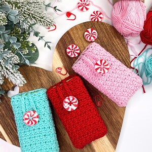 May include: Three crocheted phone cases in teal, red, and pink, each adorned with a peppermint candy button. The cases are displayed on a wooden cutting board with yarn and decorative elements, creating a festive holiday theme.