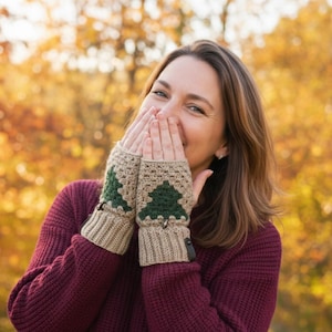 May include: Crocheted fingerless gloves with a tan base and green Christmas tree design. The gloves have a ribbed cuff and a small leather accent. The person in the image is wearing a burgundy sweater.
