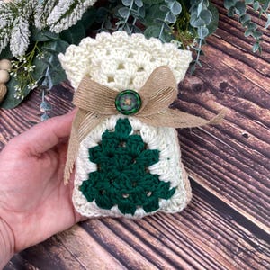 May include: A crocheted gift bag with a green Christmas tree design on the front. The bag is cream-colored with a burlap ribbon and a green button. The bag is held in a hand.
