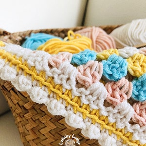 Pode incluir: Um close-up de um cobertor de crochê em branco, amarelo, rosa e azul. O cobertor está em uma cesta de vime com novelos de lã nas mesmas cores.