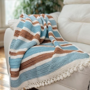 Op de afbeelding: Een handgehaakte deken met afwisselende strepen in lichtblauw, bruin en wit. De deken is over een witte fauteuil gedrapeerd. De rand van de deken heeft een witte pompom-afwerking. Een gezellig woondecoratie-item.