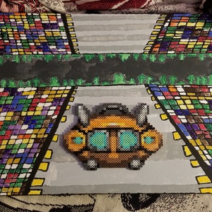 May include: A pixel art image of a yellow and brown robot with blue eyes, set against a gray background. The robot is surrounded by two colorful mosaic patterns.