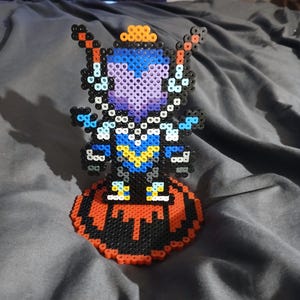 May include: A purple and blue pixel art figure with a yellow crown standing on a red and black pixel art base. The figure has a black and white design on its chest and is holding two red swords.