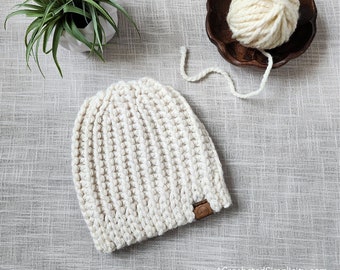Pode incluir: Um gorro de tricô branco com uma etiqueta de couro marrom. O gorro está sobre uma superfície texturizada cinza claro. Há um novelo de lã branca no fundo.
