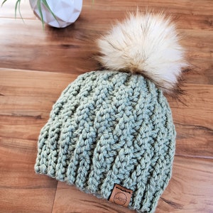 May include: A sage green, hand-crocheted beanie with a large, fluffy, cream-colored faux fur pom-pom. The hat features a textured stitch pattern and a small, brown leather tag. A small white geometric planter with a green plant is in the background.
